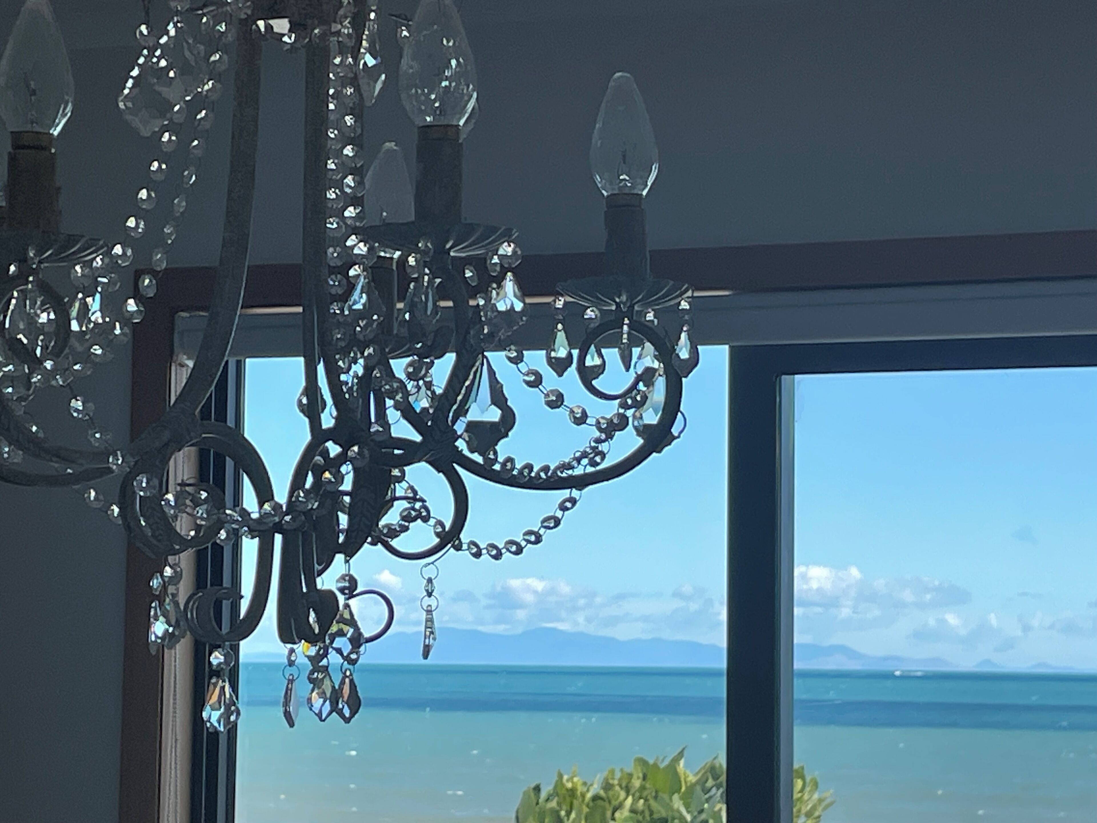 A chandelier with multiple arms and hanging crystals, a window with a view of the sea and mountains, and a glimpse of greenery outside.