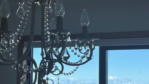 Un candelabro con múltiples brazos y cristales colgantes, una ventana que ofrece vistas al mar y a las montañas, y un atisbo de vegetación en el exterior.