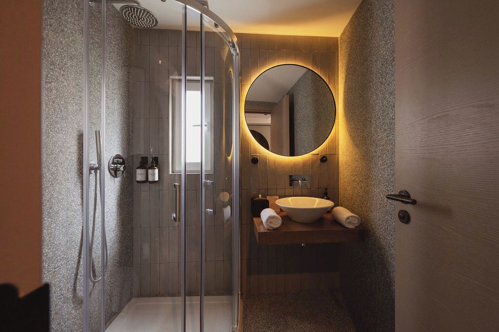 Un baño moderno con ducha de vidrio, un espejo redondo y un tocador de madera con lavamanos.
