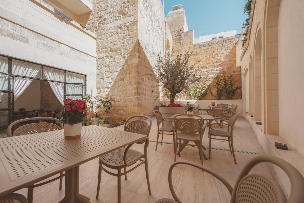 An outdoor seating area with tables and chairs, surrounded by stone walls and a potted plant with red flowers.