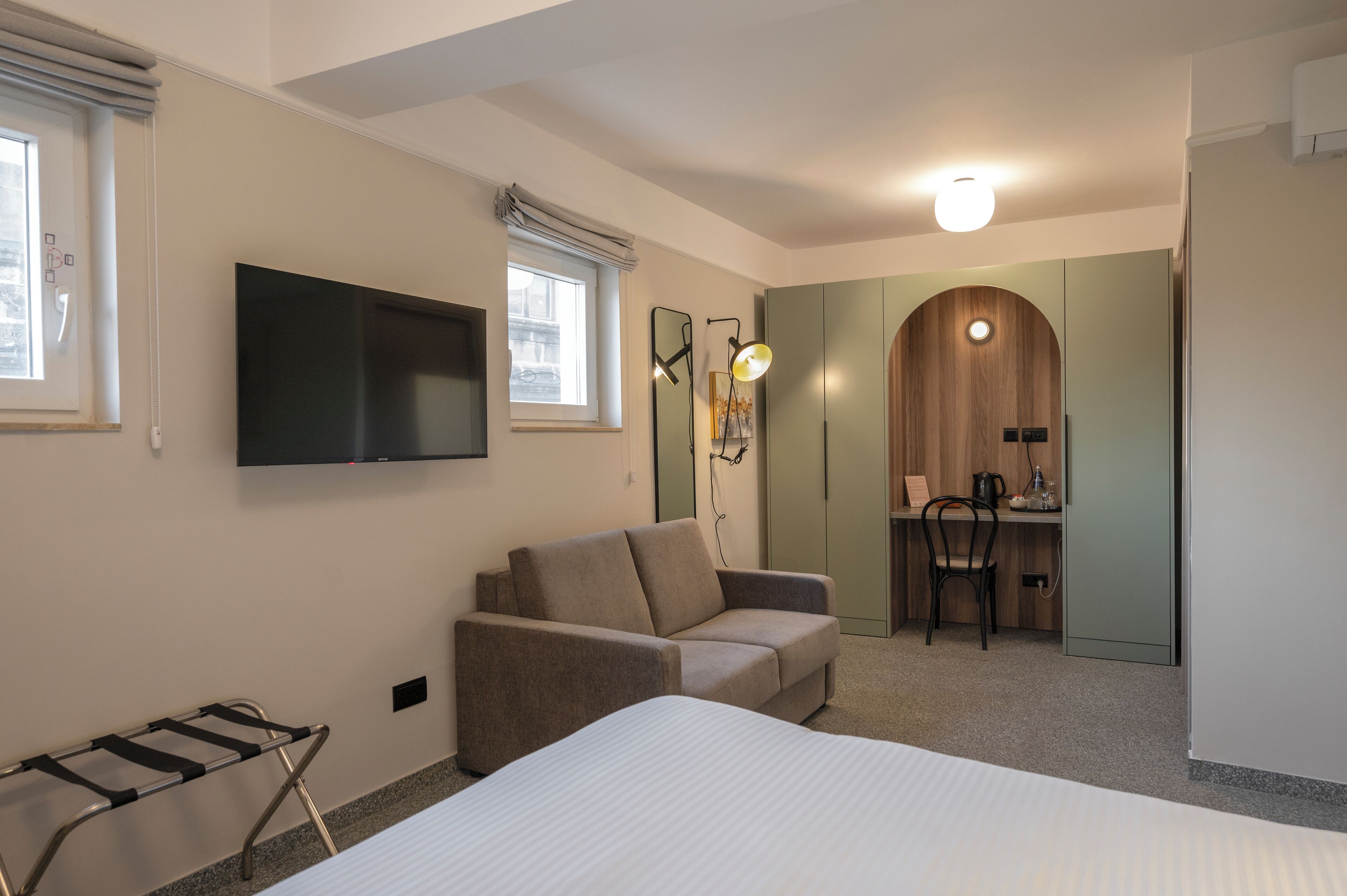 A hotel room with a bed, a sofa, a desk with a chair, a TV mounted on the wall, and a wooden door.