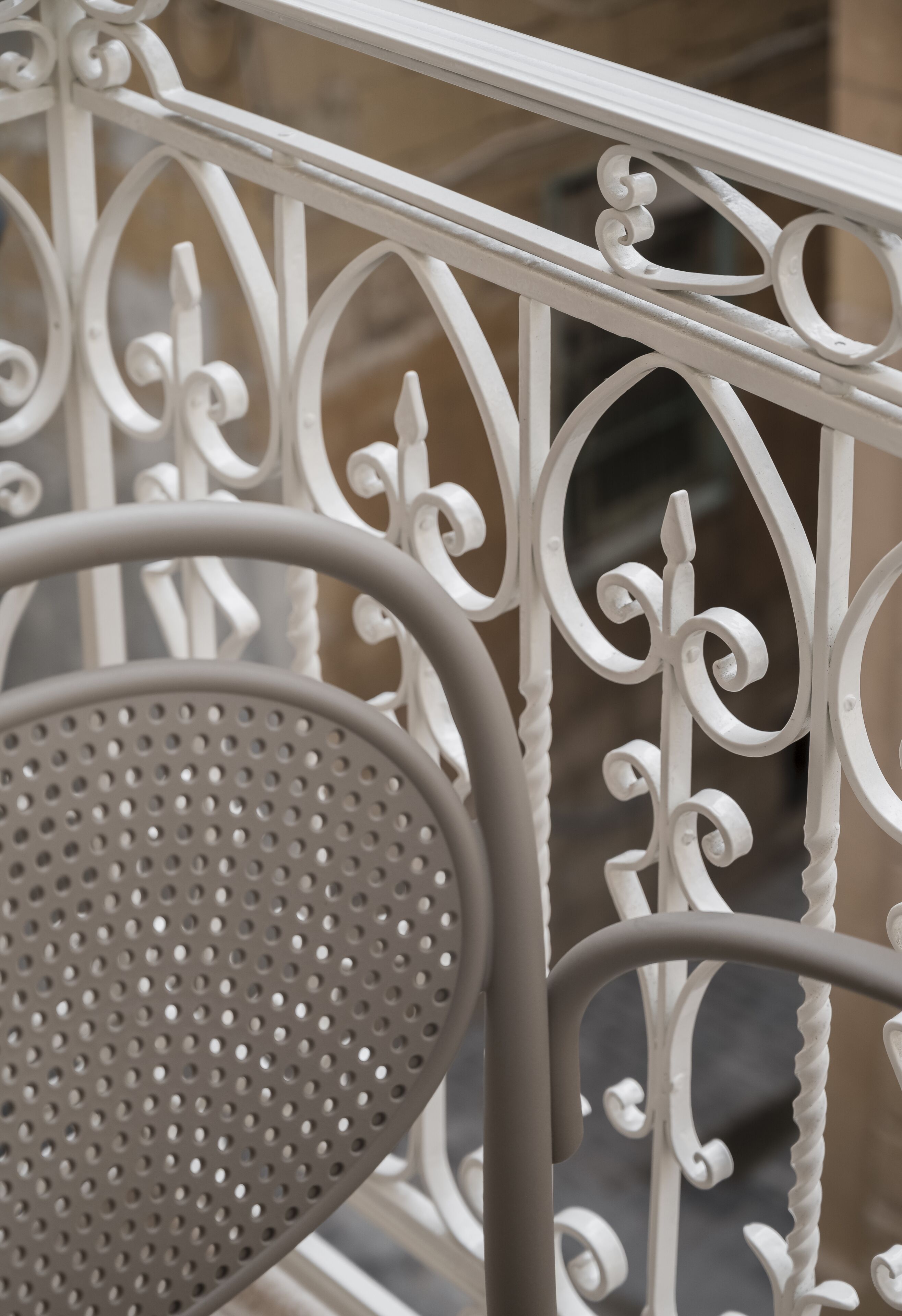 A close-up of a white metal railing with intricate designs, a perforated metal chair, and a blurred background suggesting an outdoor setting.