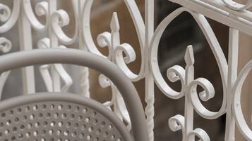 A close-up of a white metal railing with intricate designs, a perforated metal chair, and a blurred background suggesting an outdoor setting.
