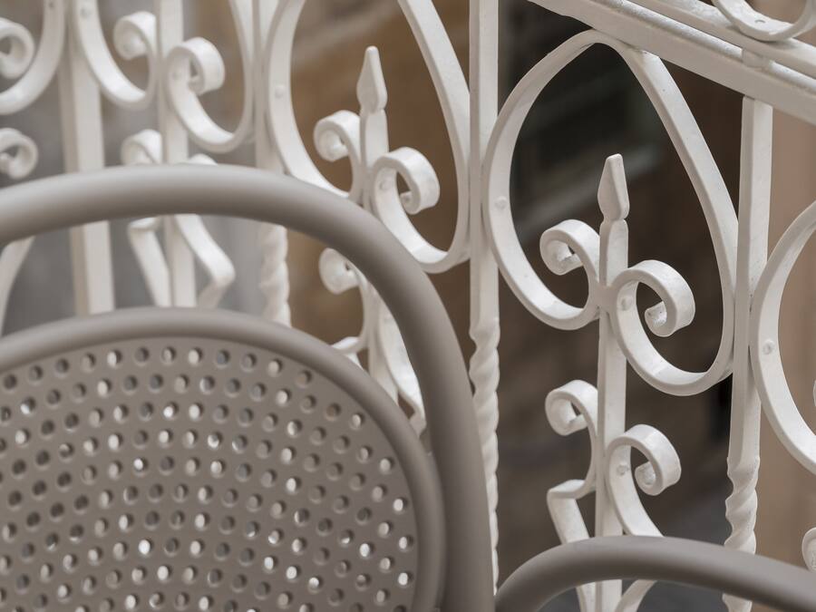 A close-up of a white metal railing with intricate designs, a perforated metal chair, and a blurred background suggesting an outdoor setting.