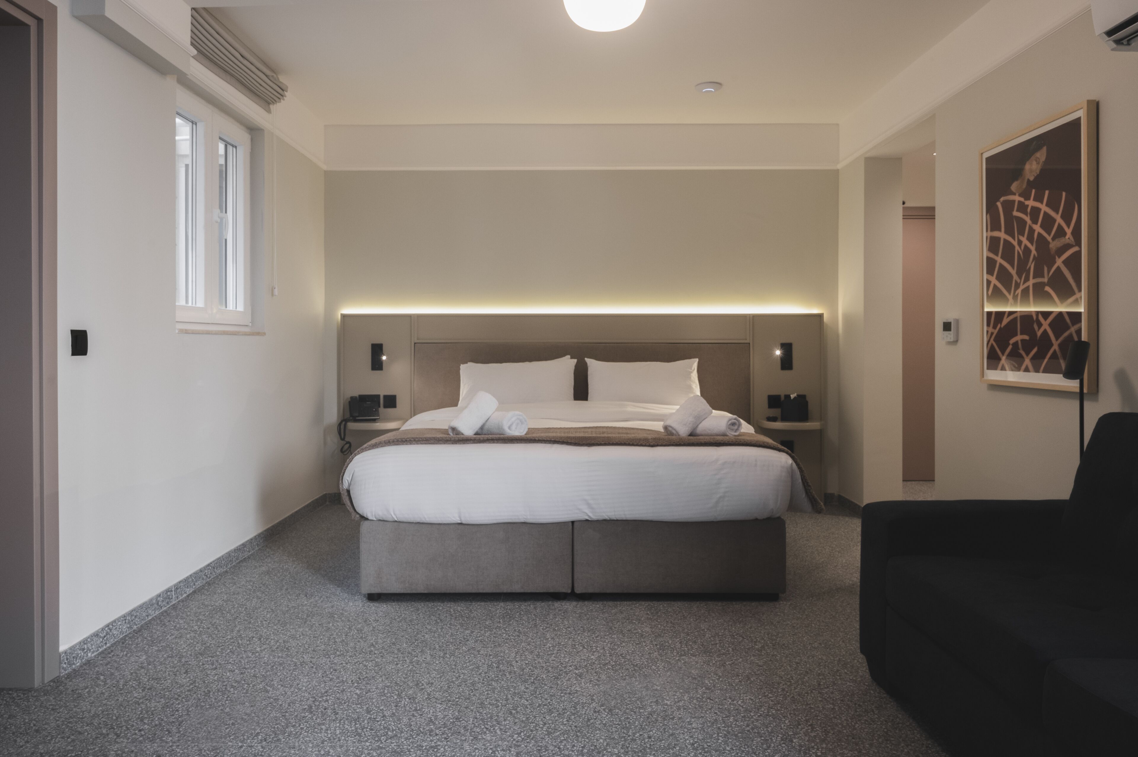 A modern hotel room with a large bed, bedside tables, a headboard with integrated lighting, a black armchair, and a large abstract painting on the wall.