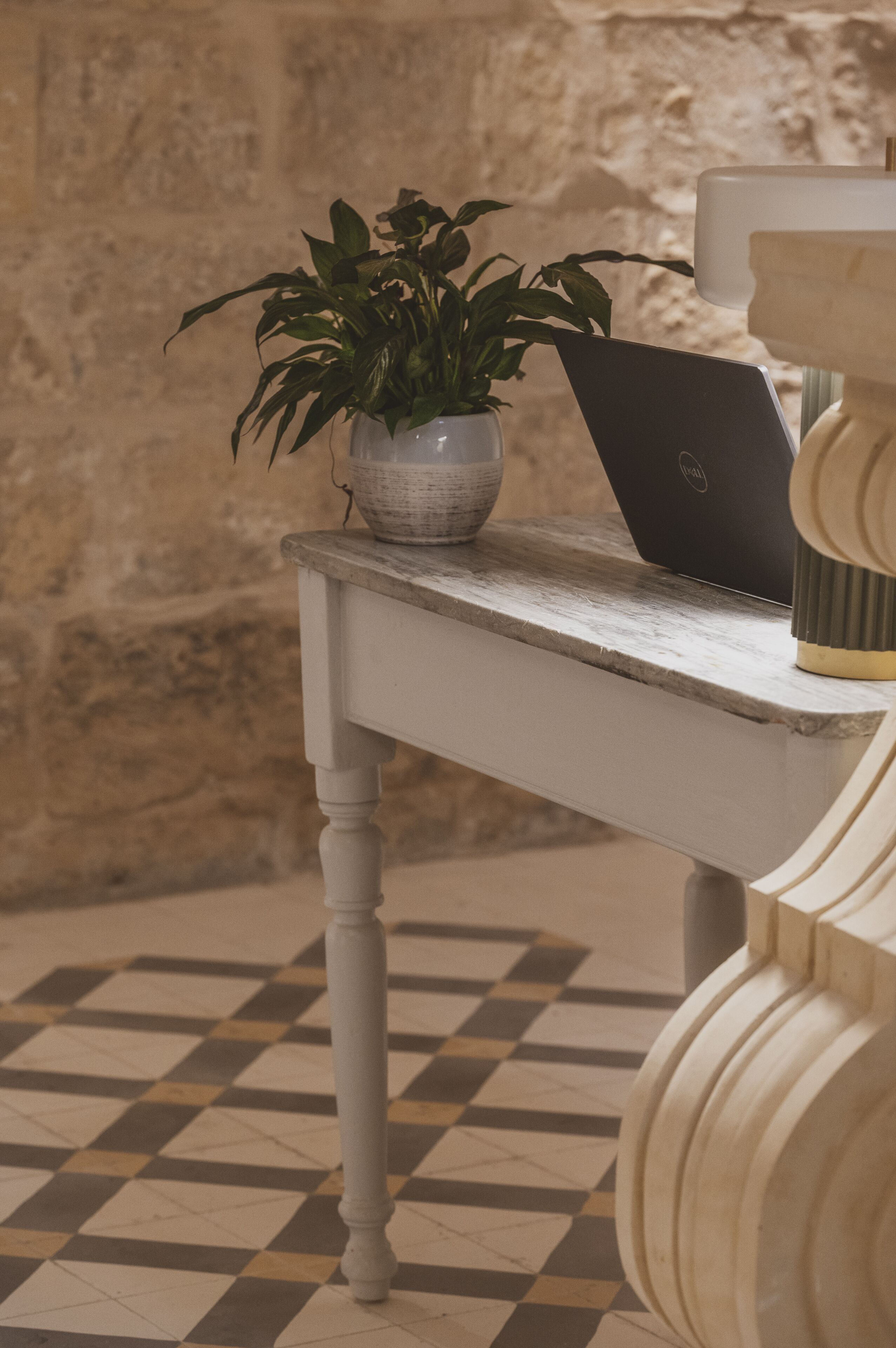 A checkered floor, a white table with a laptop and a potted plant, and a stone wall.