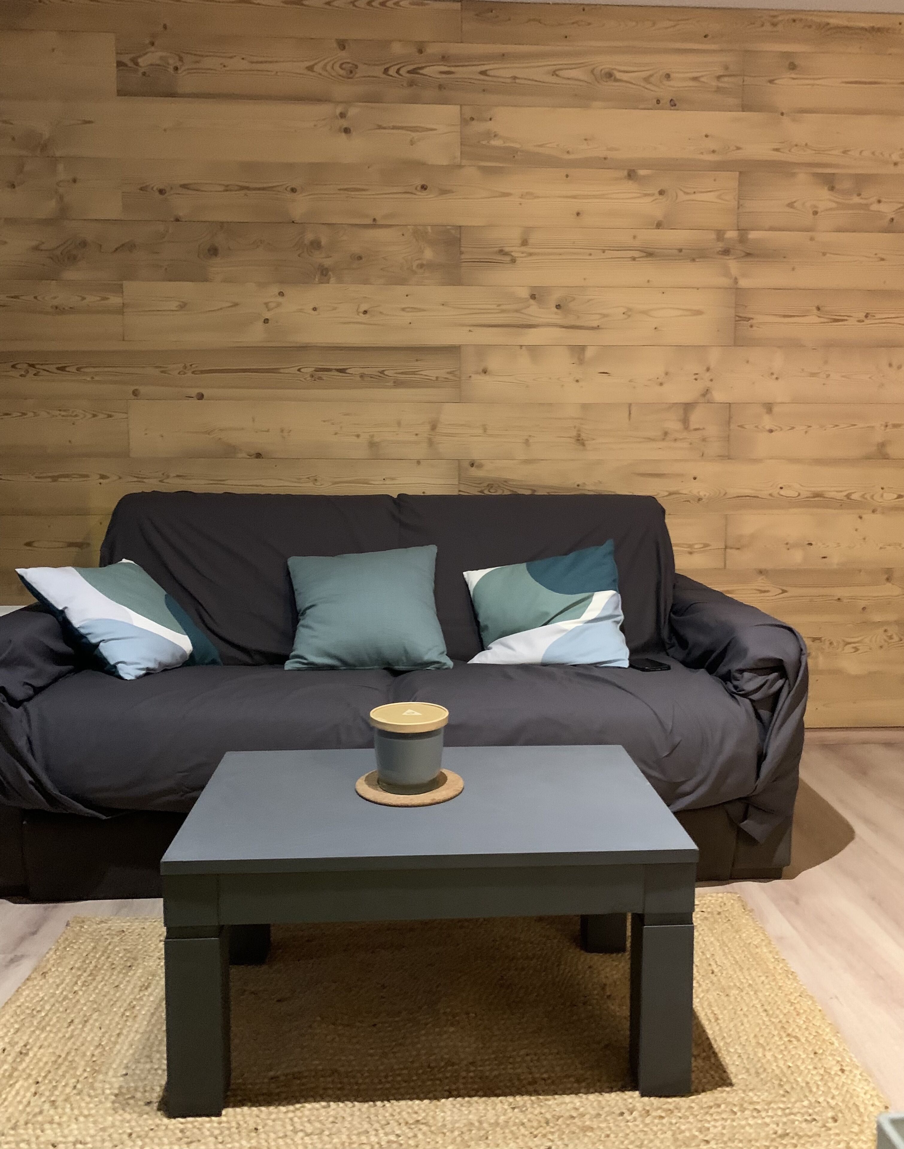 A dark-colored sofa with multiple cushions against a wooden wall, a small round table with a lid in front, and a woven rug on the floor.