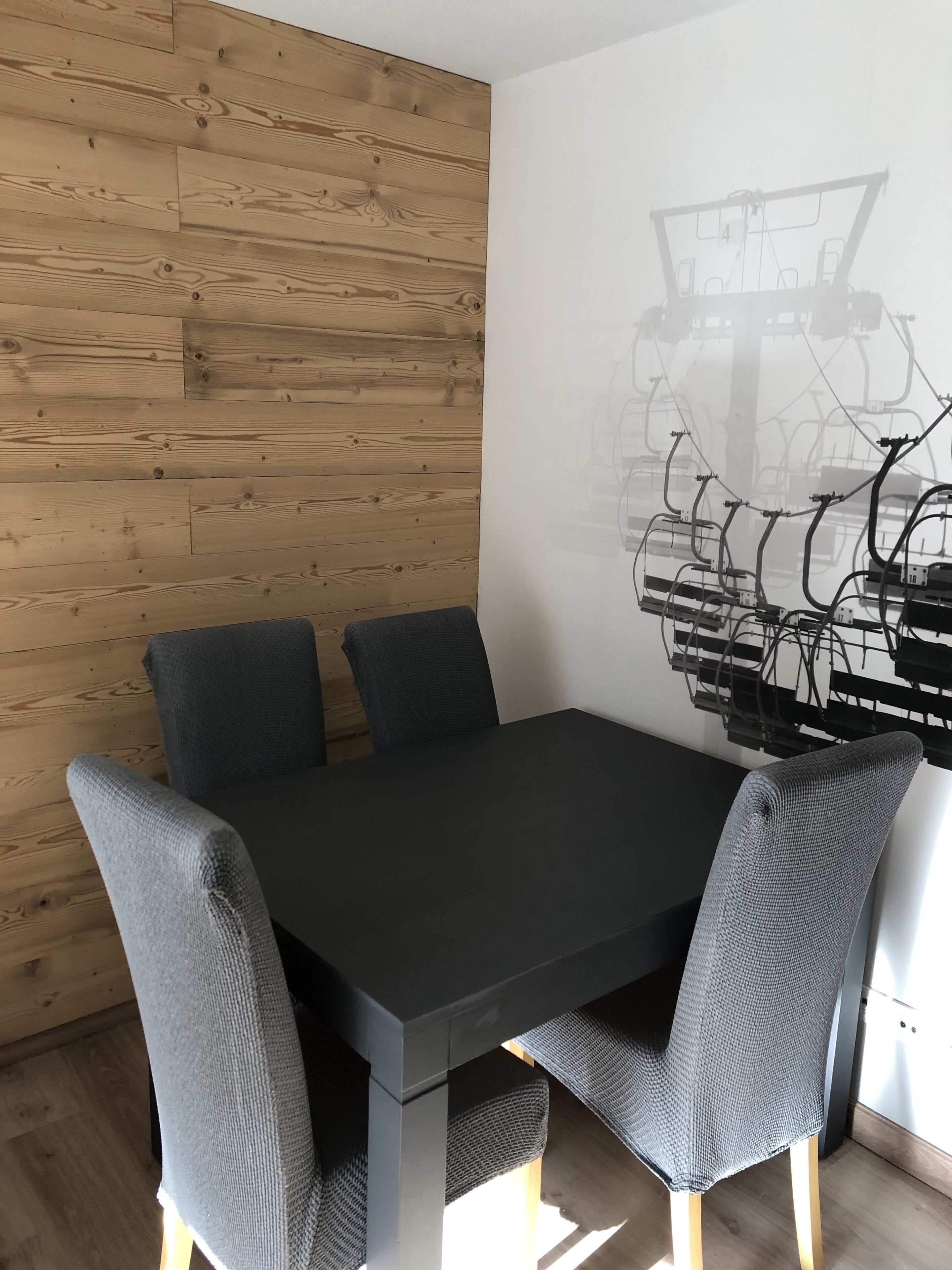 A modern dining area with a black table and grey upholstered chairs, wooden panel walls, and a white wall with a black abstract art piece.