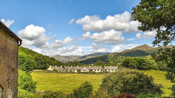 A traditional stone house with a stone wall in the foreground, set against a backdrop of lush green hills and a clear blue sky. - Church House Cottage (Ulpha)
