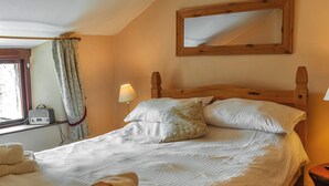 A neatly made bed with pillows and a blanket, a bedside lamp, a wooden headboard, a window with curtains, and a wall-mounted mirror. - Church House Cottage (Ulpha)