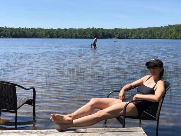 Une personne assise sur une chaise longue au bord d’un lac, avec un bateau en arrière-plan.
