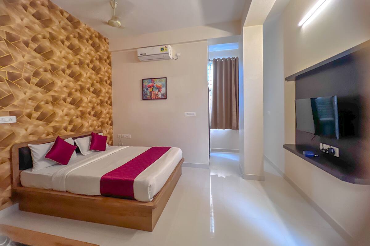 Une chambre d’hôtel moderne avec une tête de lit en bois, un grand lit avec une literie blanche et un plaid rouge, un téléviseur à écran plat fixé au mur et un ventilateur de plafond.