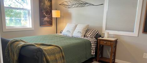 A bedroom with a bed, a nightstand, a lamp, a window, and a painting on the wall.