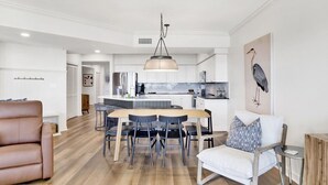 Una sala moderna con un sofá de cuero marrón, un sillón blanco y una mesa de comedor con sillas. Se ve una cocina al fondo con armarios blancos y una lámpara colgante.