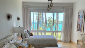Ein Hotelzimmer mit zwei Betten, einem großen Fenster mit Blick auf Palmen und das Meer sowie einem Gemälde an der Wand.