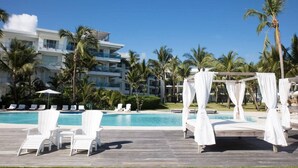Ein Resort-Poolbereich mit weißen Liegestühlen, einem Baldachinbett und freiem Blick auf ein mehrstöckiges Gebäude im Hintergrund.