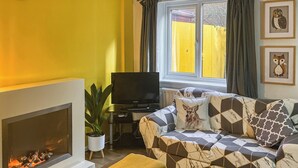 A living room with a fireplace, a sofa with patterned cushions, a television, and a window with curtains.
