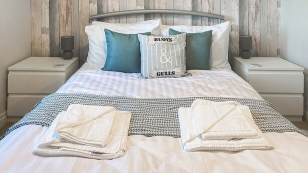 A neatly made bed with a striped blanket, two pillows with a nautical design, and a bedside table with a lamp.