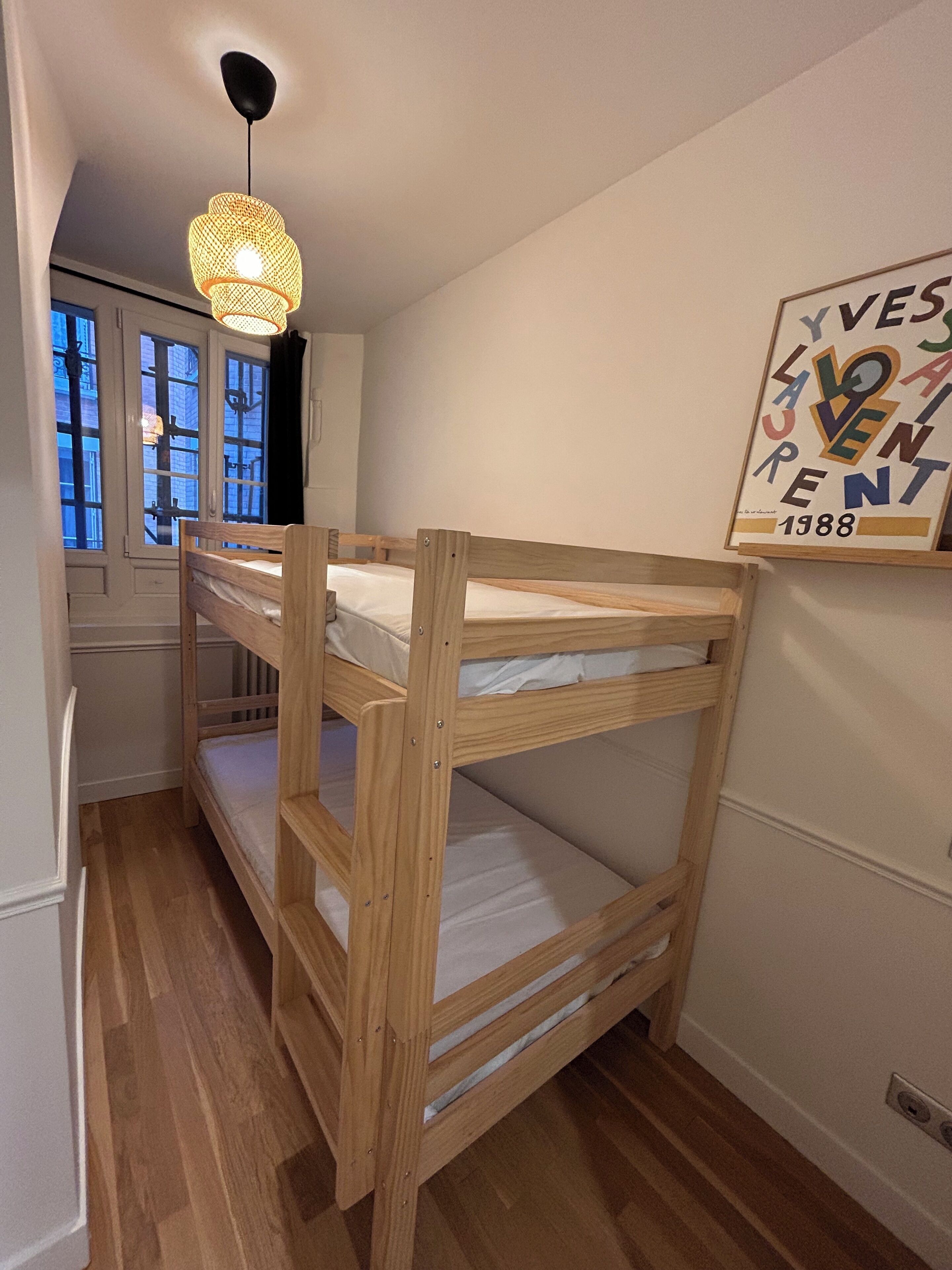 A bunk bed room with a wooden floor, a window with curtains, and a hanging light fixture.