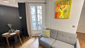 Ein Wohnzimmer mit grauem Sofa, einem Esstisch mit Stühlen, einer Kristallleuchtre und einem Fenster mit Blick auf Gebäude.