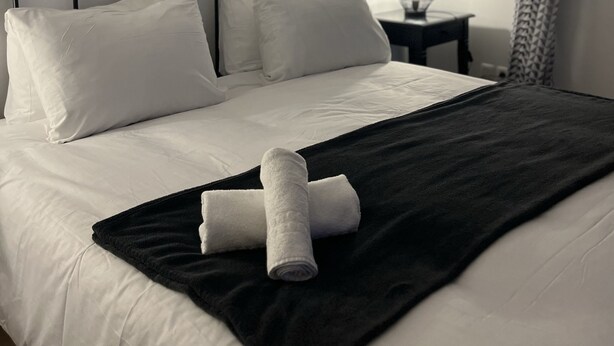 Una cama bien tendida con sábanas blancas y un plaid negro, una mesita de noche con lámpara, y una ventana con cortina estampada.