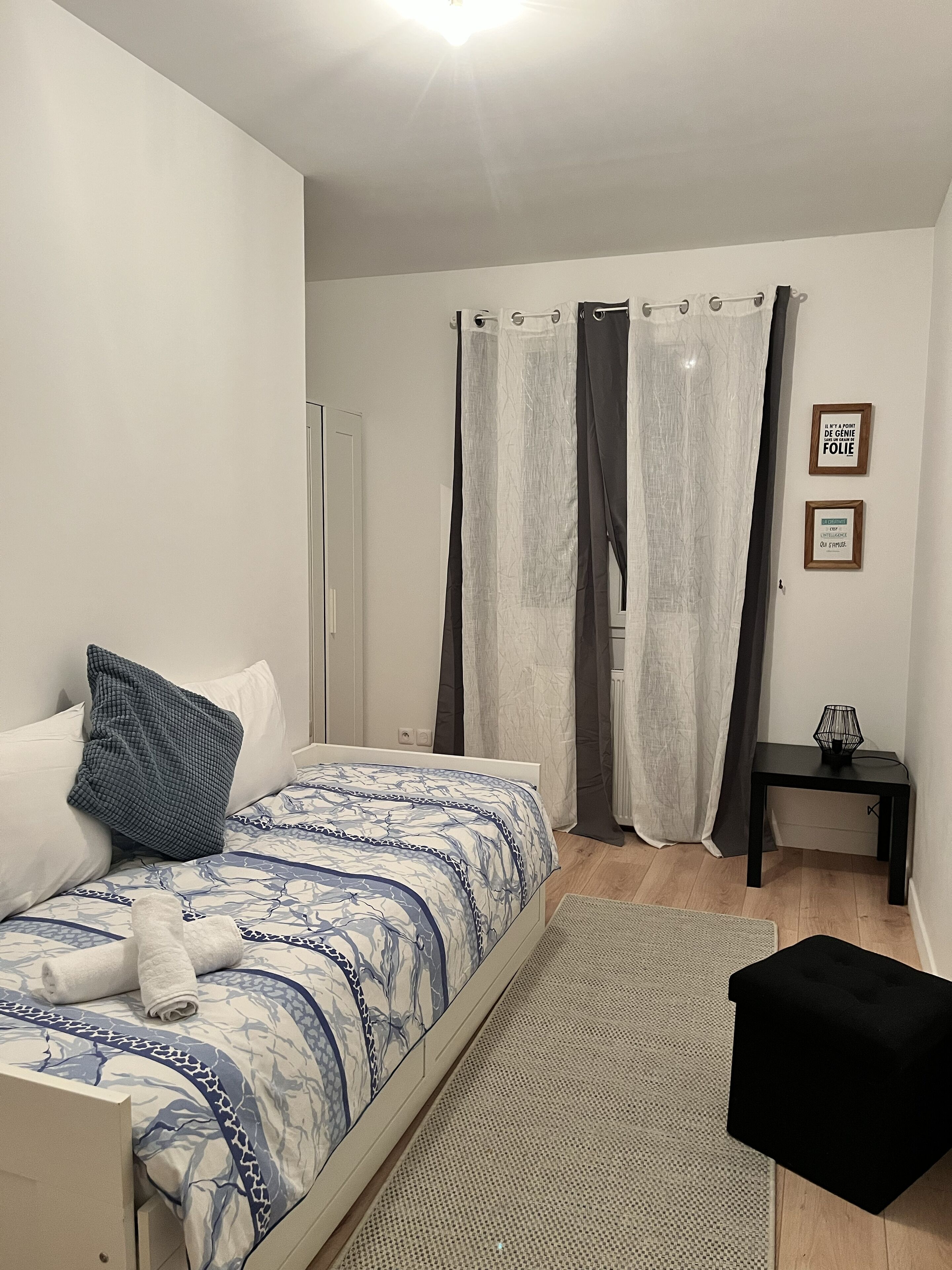 A bedroom with a bed, a nightstand, a black ottoman, a rug, and two framed quotes on the wall.