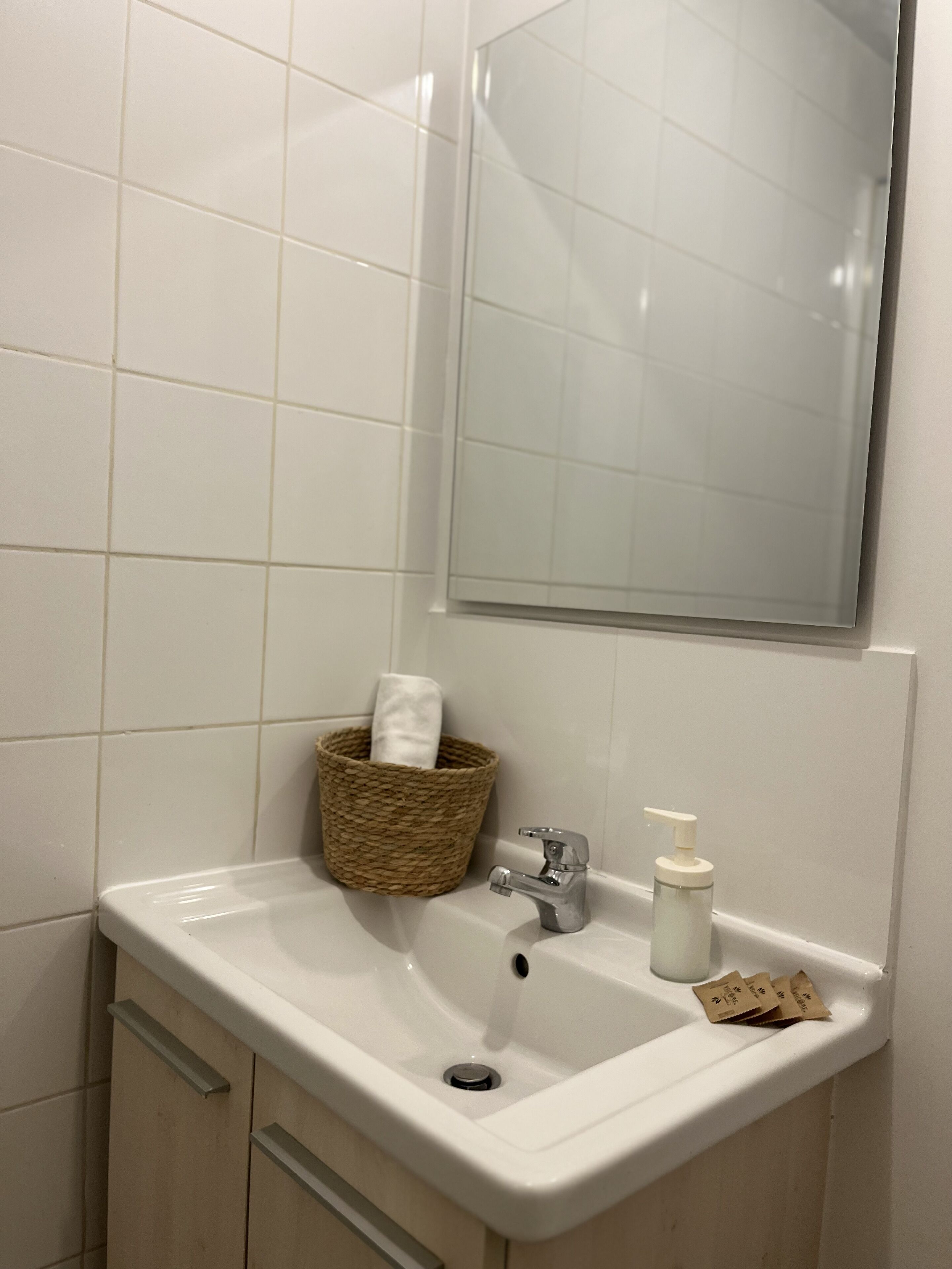 A bathroom with a white sink, a mirror, a wicker basket with a towel, and a soap dispenser.