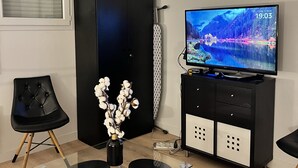 Una sala de estar moderna con una mesa de comedor de vidrio, sillas negras, un mueble de TV negro y un jarrón con flores blancas.