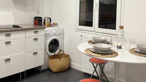 Una cocina compacta con armarios blancos, lavadora, mesa de comedor con taburetes y ventana con vistas.