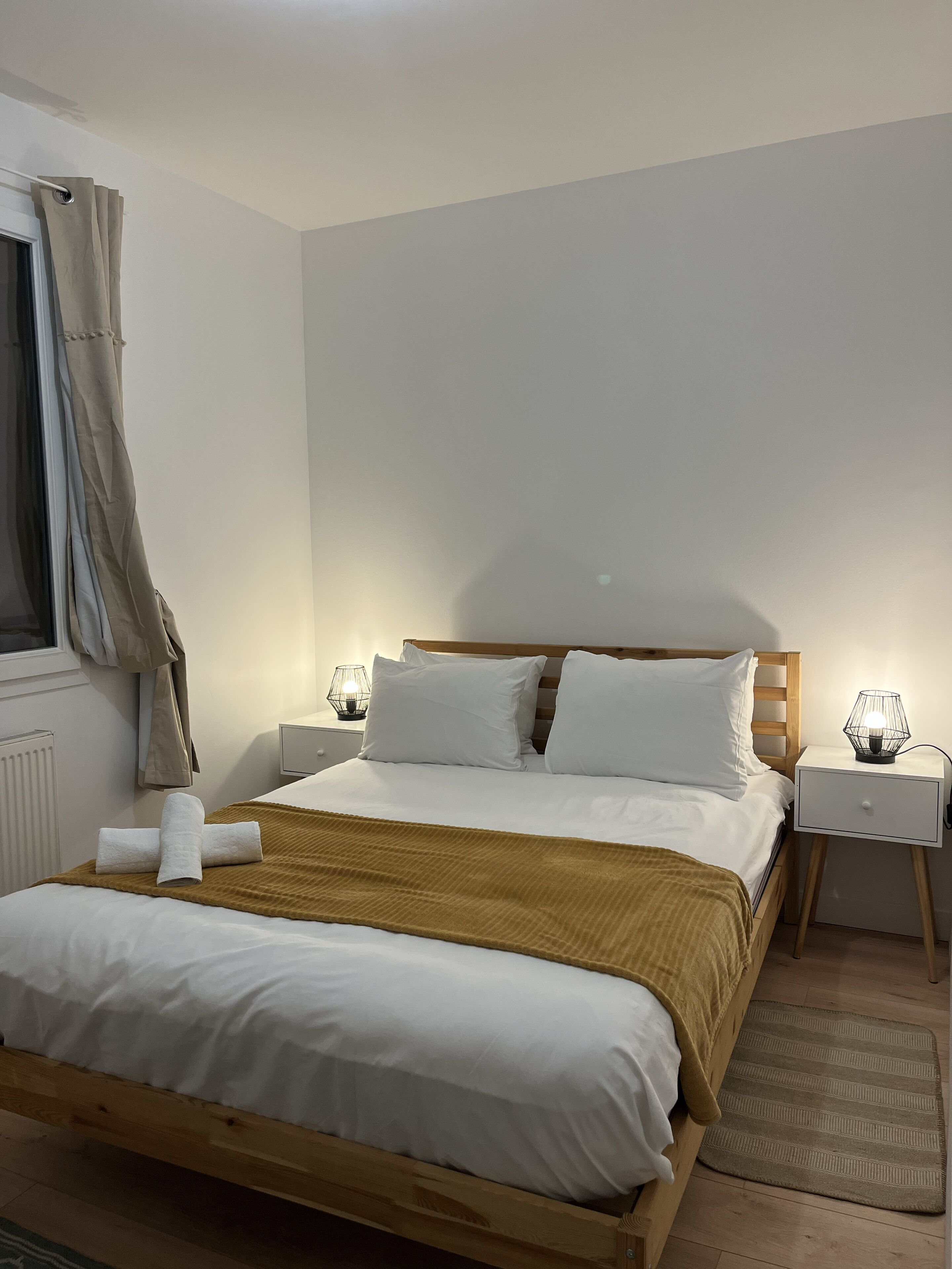 Un lit bien fait, avec du linge de lit blanc et un plaid marron, une tête de lit en bois, deux tables de chevet avec des lampes et une fenêtre avec des rideaux.