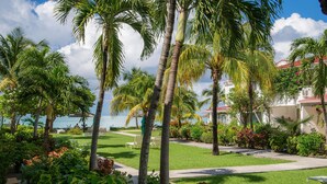 A tropical garden with palm trees, a pool, and a view of the sea. - Romantic Antigua Getaway – Oceanfront Condo with Resort Amenities (St. John's)