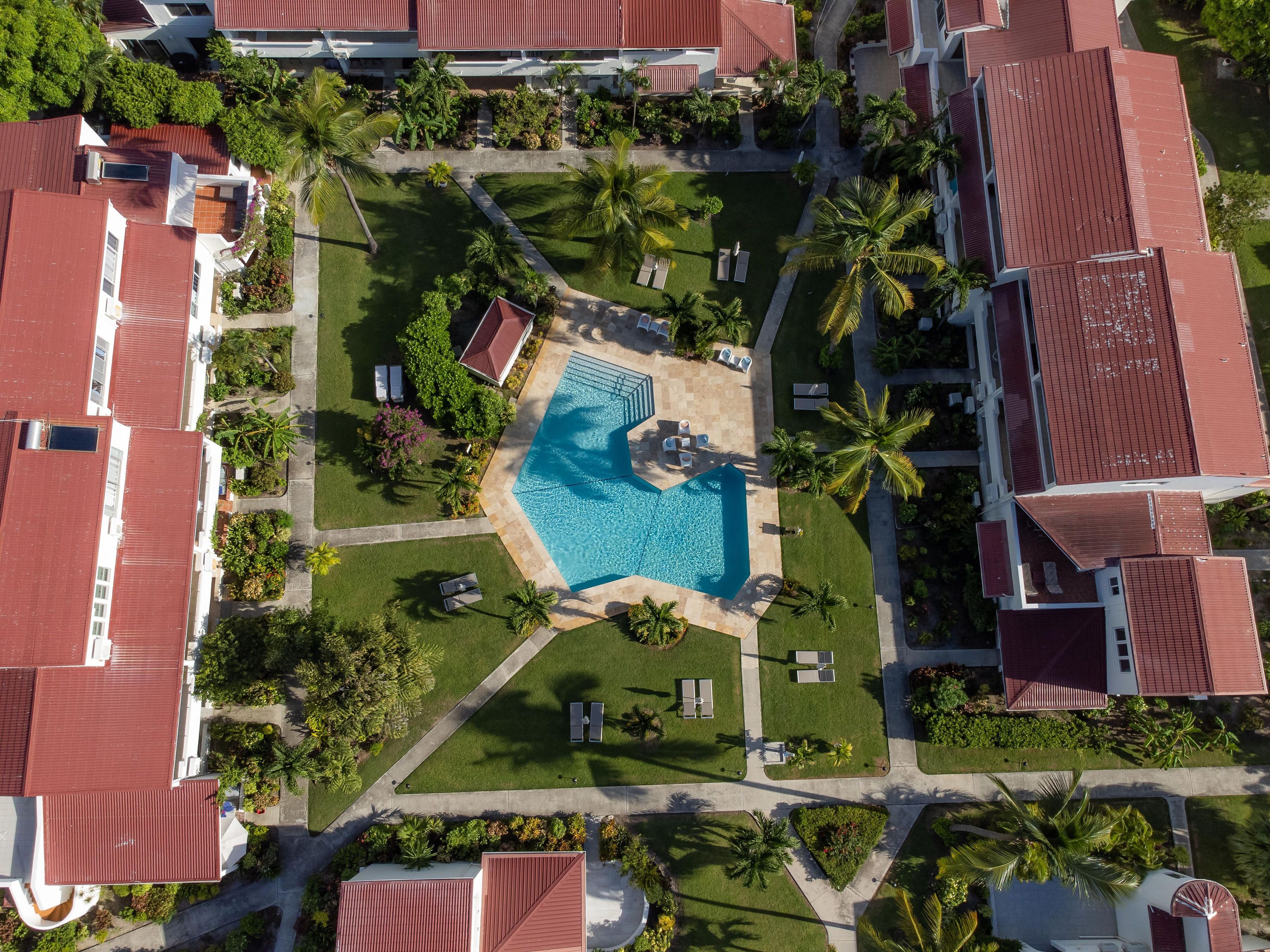 An aerial view of a resort with a central swimming pool, surrounded by lush greenery and palm trees.