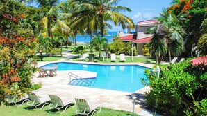 A swimming pool surrounded by palm trees and lounge chairs, with a view of the ocean in the background. - Romantic Antigua Getaway – Oceanfront Condo with Resort Amenities (St. John's)