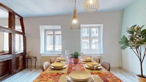 A dining area with a table set for six, a potted plant, and a view of buildings through windows. - Le petit chez vous (Colmar)