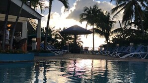 A pool area with lounge chairs and umbrellas, surrounded by palm trees and a building with a covered seating area. - Large Pool/ 1 BR/1Bath  Apt. in Beautiful St. Martin in Simpson Bay! (Simpson Bay)