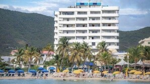 A multi-story hotel with balconies, located near a beach with palm trees and beach umbrellas. - Large Pool/ 1 BR/1Bath  Apt. in Beautiful St. Martin in Simpson Bay! (Simpson Bay)