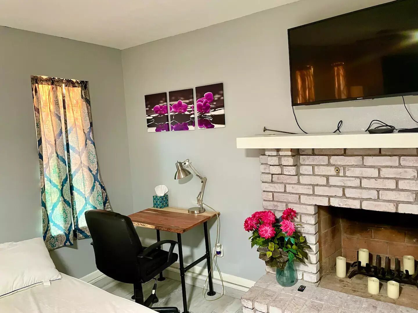 Quarto com cama, escrivaninha com cadeira, televisão de tela plana acima de lareira e janela com cortinas.