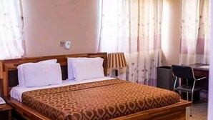 A neatly made bed with a patterned bedspread, a bedside lamp, and a chair in a room with curtains and a desk. - Whyte View Lodge (Sekondi-Takoradi)