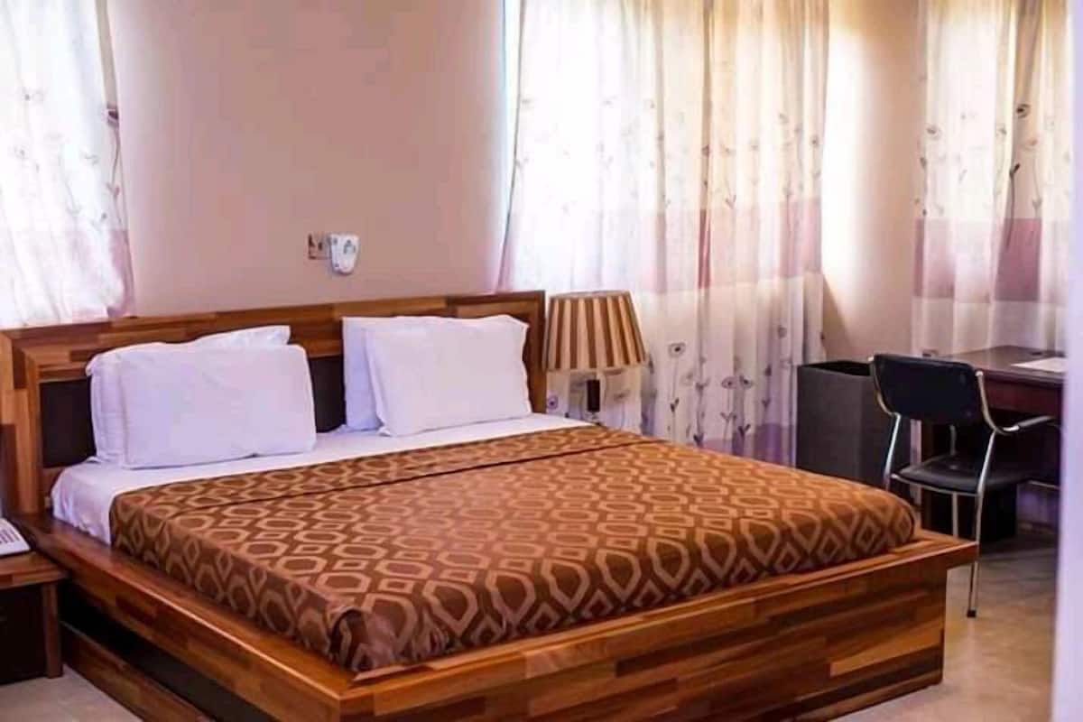 A neatly made bed with a patterned bedspread, a bedside lamp, and a chair in a room with curtains and a desk.