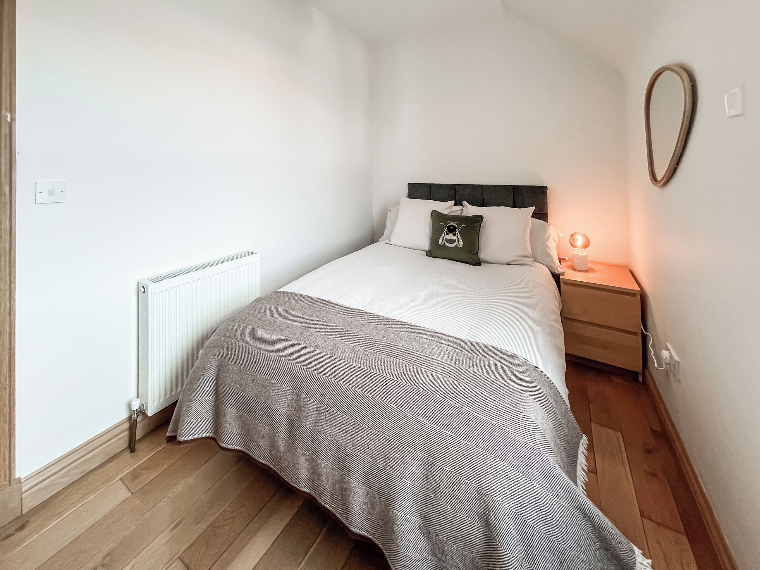 A bedroom with a bed, a nightstand, a bedside lamp, a radiator, and a wooden floor.