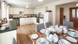 Una cocina moderna con armarios blancos, una zona de comedor con mesa de madera dispuesta para seis personas, y un pequeño rincón de estar con sillas.