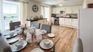Un comedor con una mesa de madera lista para una comida, una cocina con alacenas blancas y un reloj grande en la pared.