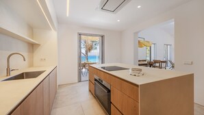 A modern kitchen with a large island, built-in oven, and a view of the ocean through a glass door. - VILLA ES BARCARES (Alcúdia)