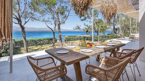 An outdoor dining area with a view of the sea, wicker furniture, and a wooden table set for two. - VILLA ES BARCARES (Alcúdia)