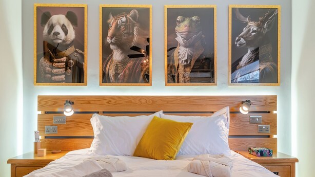 Un lit bien fait, avec du linge de lit blanc et gris, un coussin jaune et une tête de lit en bois agrémentée de lampes fixées au mur.