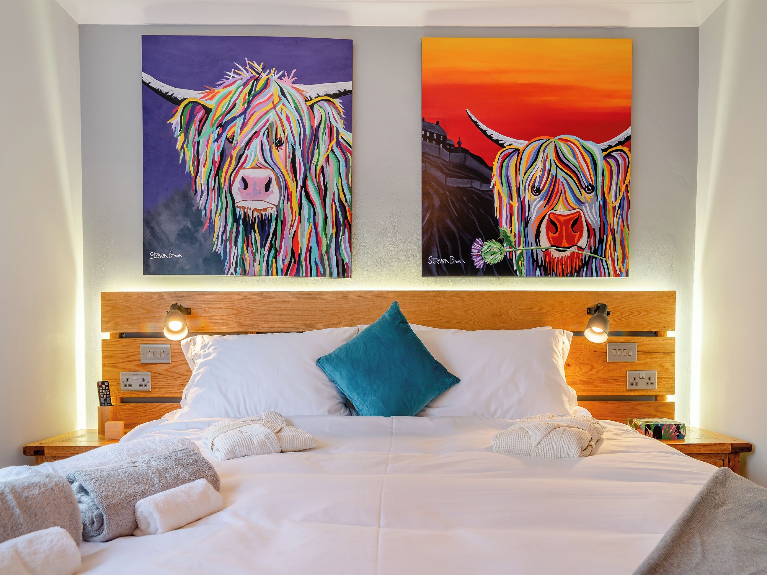 A hotel room with a large bed, two paintings of bulls, a wooden headboard, bedside lamps, and a nightstand with a remote control.