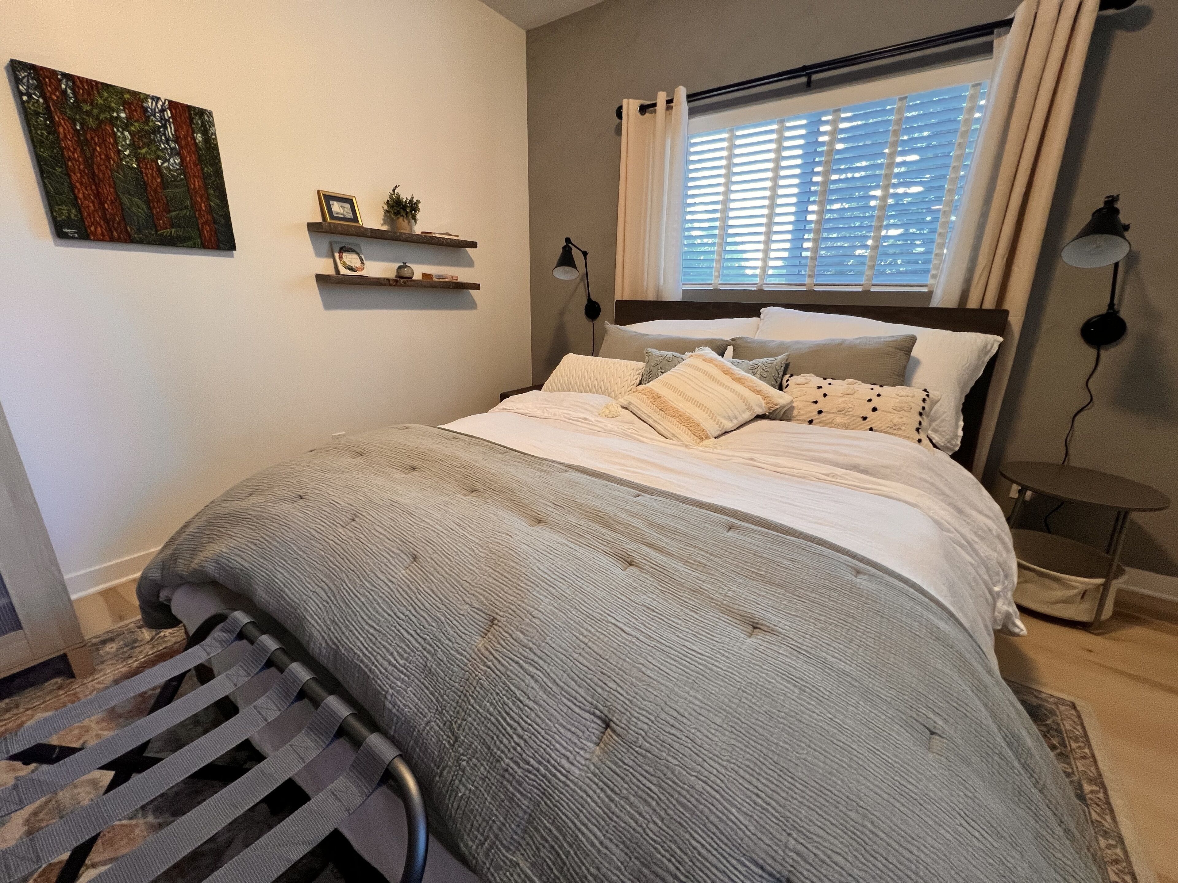 A bedroom with a bed, a window with blinds, a bedside lamp, and a small shelf with decorative items.