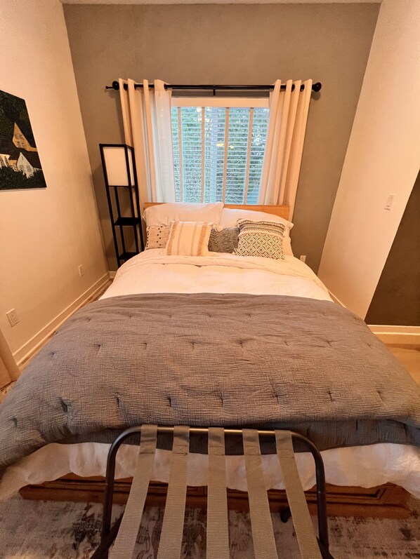 A bedroom with a bed, a window with curtains, a bedside table, and a wall-mounted shelf.