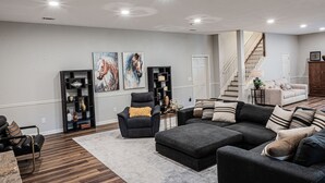 A spacious living room with a large sectional sofa, a black armchair, and a bookshelf. A staircase is visible in the background leading to another level. - Mid-Century Modern gem in the heart of the country, close to Lake Lanier.  (Auburn)