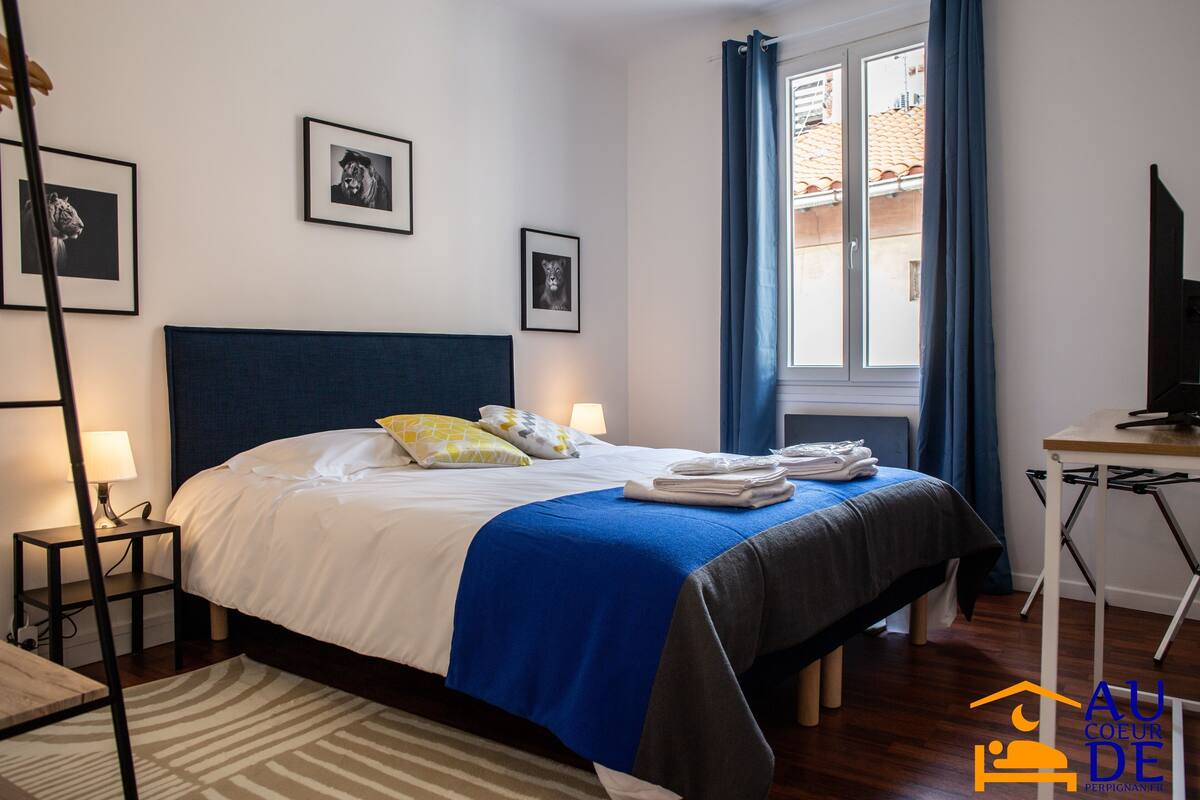A neatly made bed with a blue and white bedspread, a bedside table with a lamp, and framed pictures on the wall.